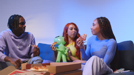 Gruppe-Von-Gen-Z-Freunden,-Die-Zu-Hause-Auf-Dem-Sofa-Sitzen,-Pizza-Zum-Mitnehmen-Essen-Und-Mit-Spielzeug-Alien-Spielen